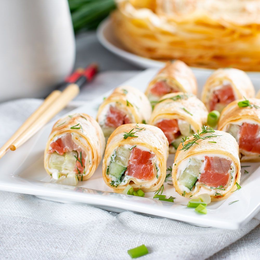Rollen mit Lachs, Frischkäse und Gurke auf einem weißen Teller. Dazu Stäbchen und eine weiße Schale.