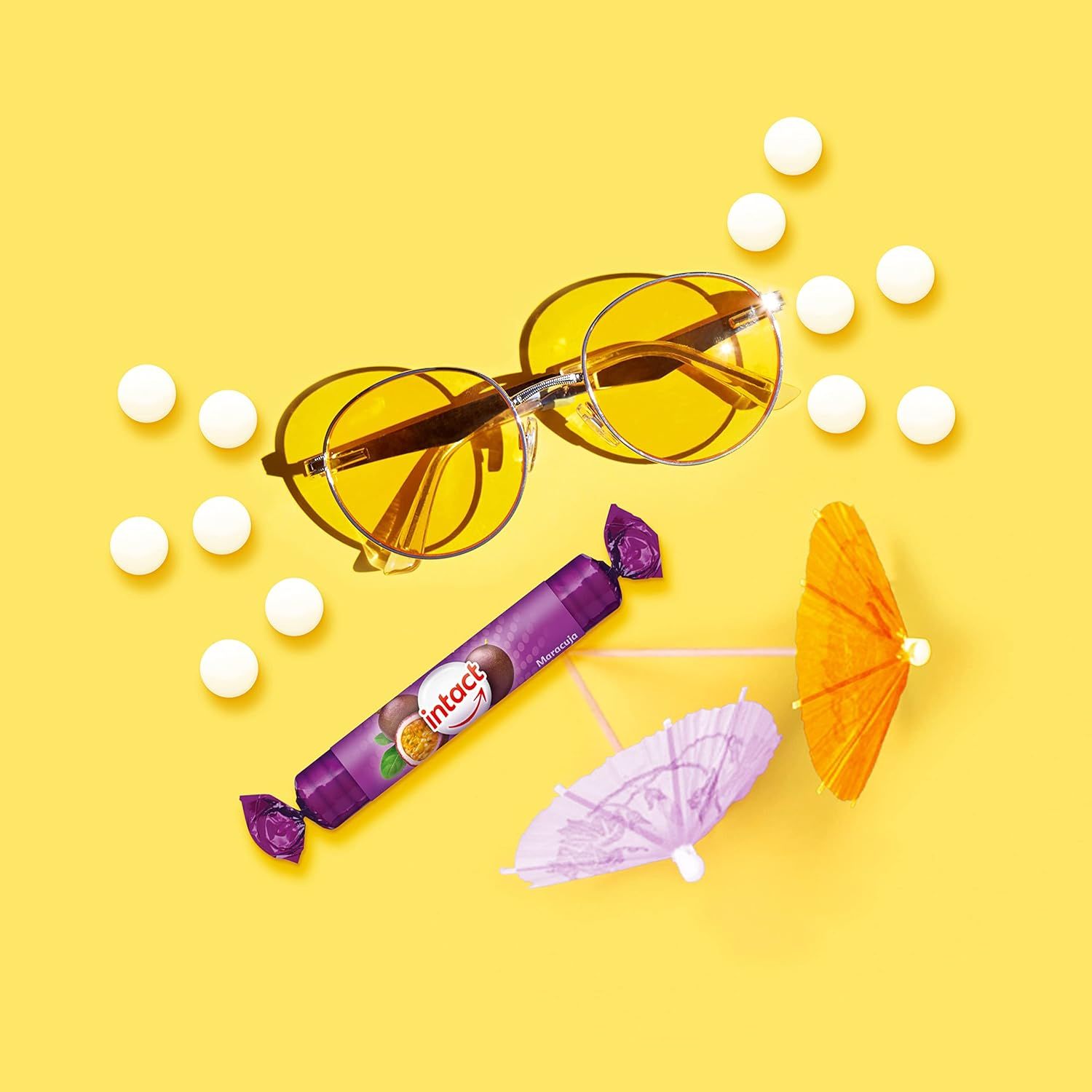 Eine Rolle Traubenzucker mit Maracuja-Geschmack, Sonnenbrille und Sonnenschirmen auf gelbem Hintergrund. Daneben weiße Tabletten.