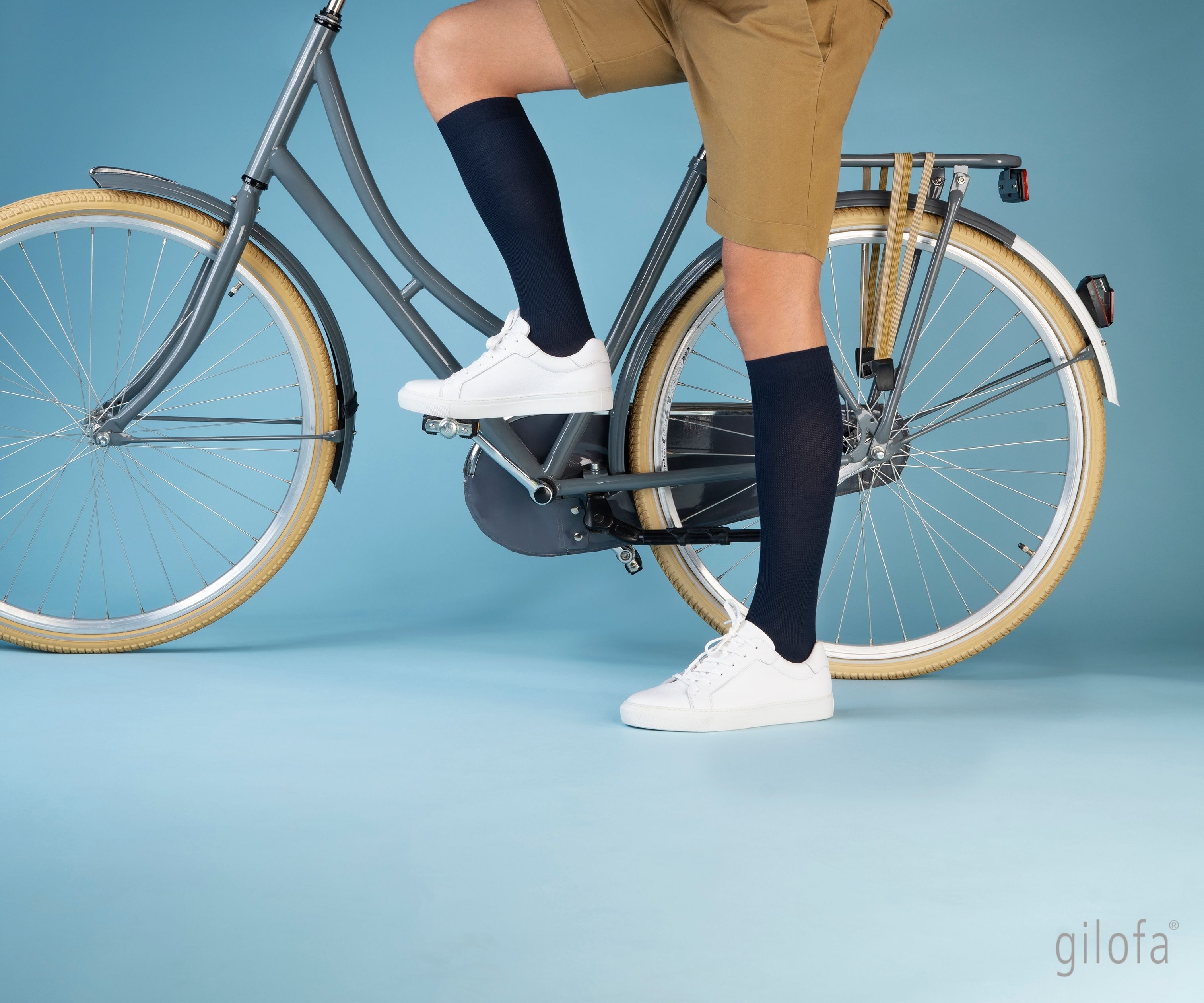Person auf einem Fahrrad mit Kniestrümpfen. Dunkelblaue Kniestrümpfe. Weiße Schuhe. Blaues Hintergrund.