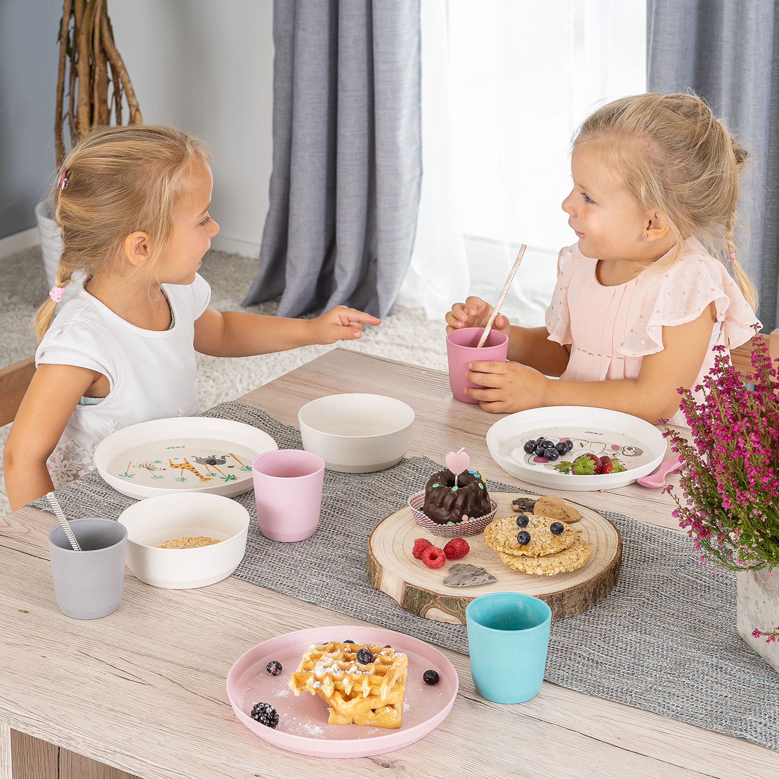 Zwei Mädchen sitzen an einem Tisch mit Kindergeschirr. Teller mit Waffeln, Beeren und Kuchen. Rosa und blaue Becher.