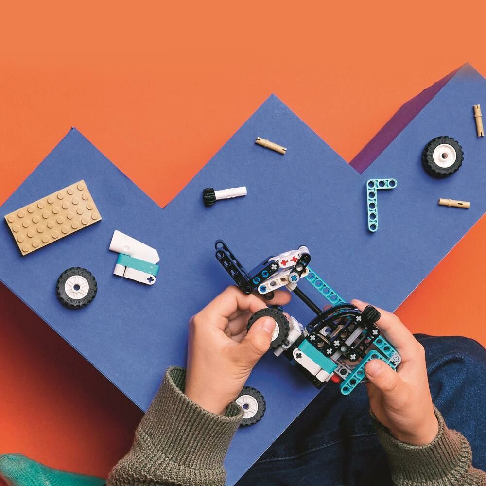 Primo piano di mani che costruiscono un modello di sollevatore telescopico LEGO Technic. Singoli pezzi blu, neri, bianchi e beige sono posizionati su una superficie blu. Il set include ruote, un braccio telescopico e una forcella.