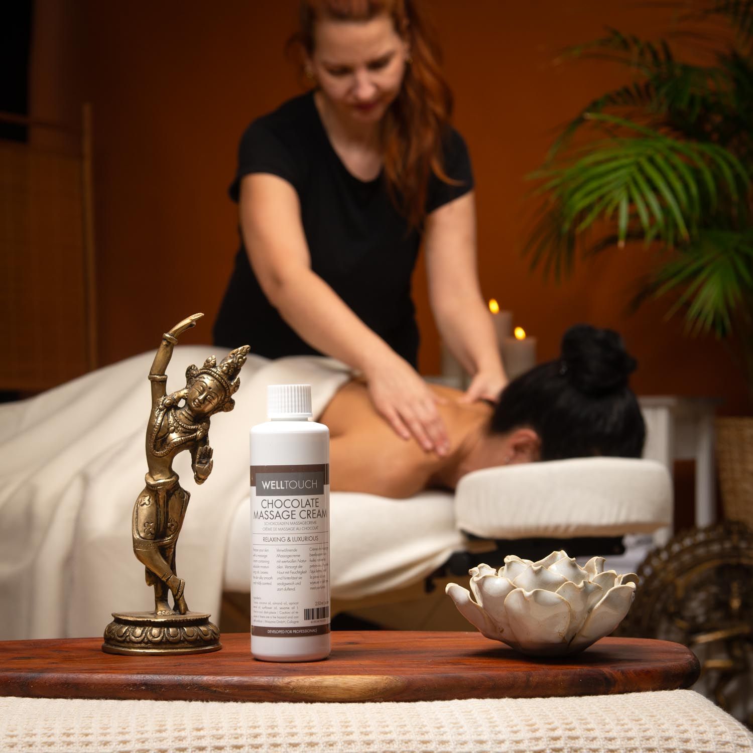 Weiße Flasche Welltouch Schokoladenmassage-Creme. Aufschrift: Chocolate Massage Cream. Brauner Deckel, weißes Etikett. Frau erhält Massage.