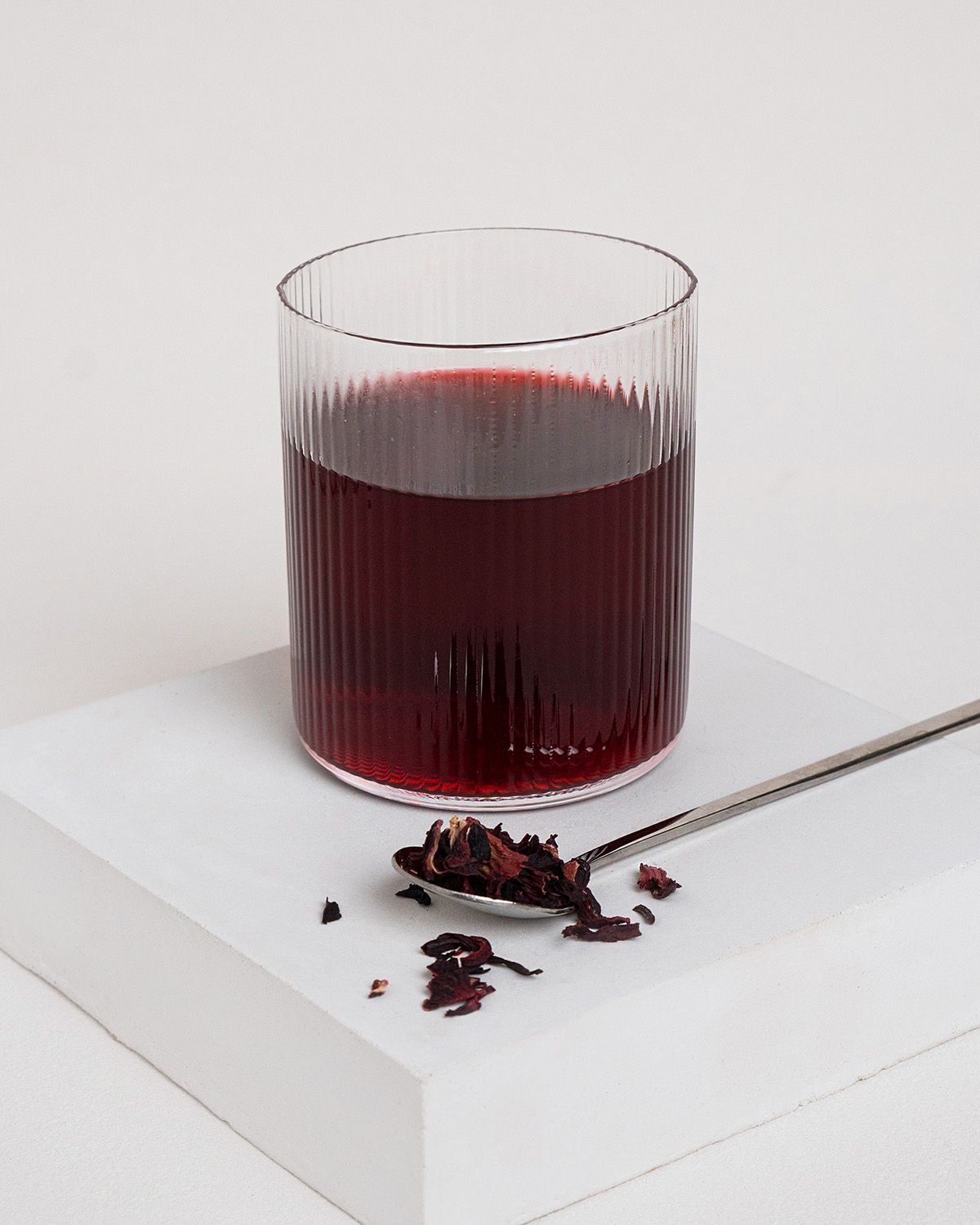Glas mit rotem Getränk und Löffel mit Hibiskusblüten. Glas mit vertikalen Rillen. Auf weißem Podest.