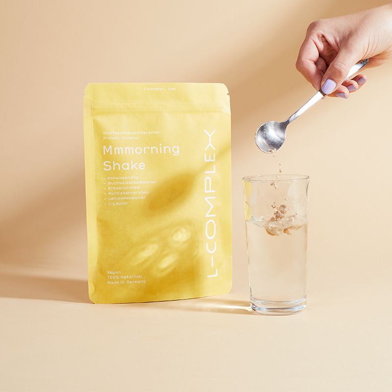 Gelbe Verpackung "Mmmorning Shake" und "L-COMPLEX" neben Glas mit Wasser. Hand mit Löffel, der Pulver in das Glas gibt.