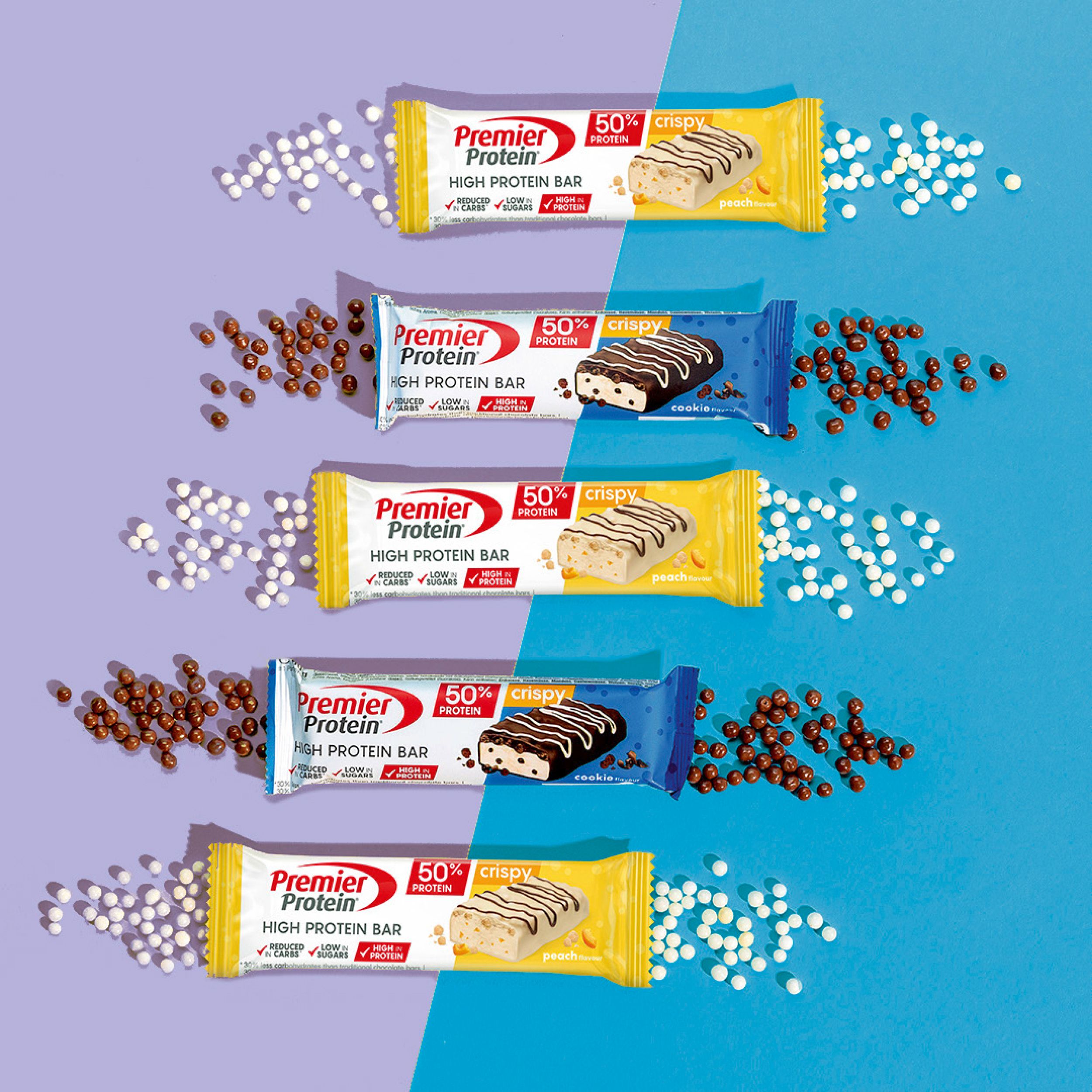 PREMIER PROTEIN High Protein Riegel. Mehrere Riegel, verschiedene Geschmacksrichtungen, umgeben von weißen und braunen Kügelchen.