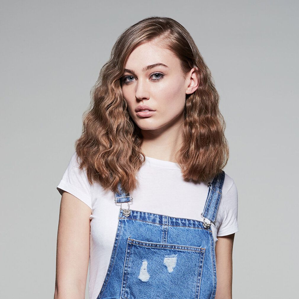 Frau mit gewelltem Haar. Sie trägt ein weißes T-Shirt und eine Jeans-Latzhose. Grauer Hintergrund.