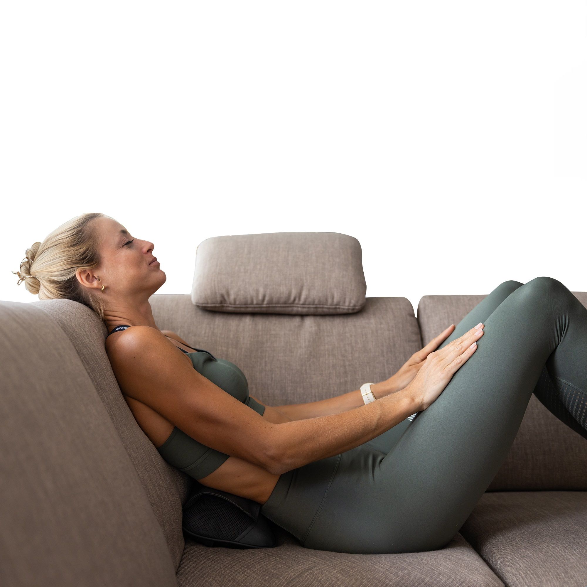 Frau liegt auf Sofa, Massagekissen am unteren Rücken. Schwarzes Kissen. Frau in Sportkleidung.