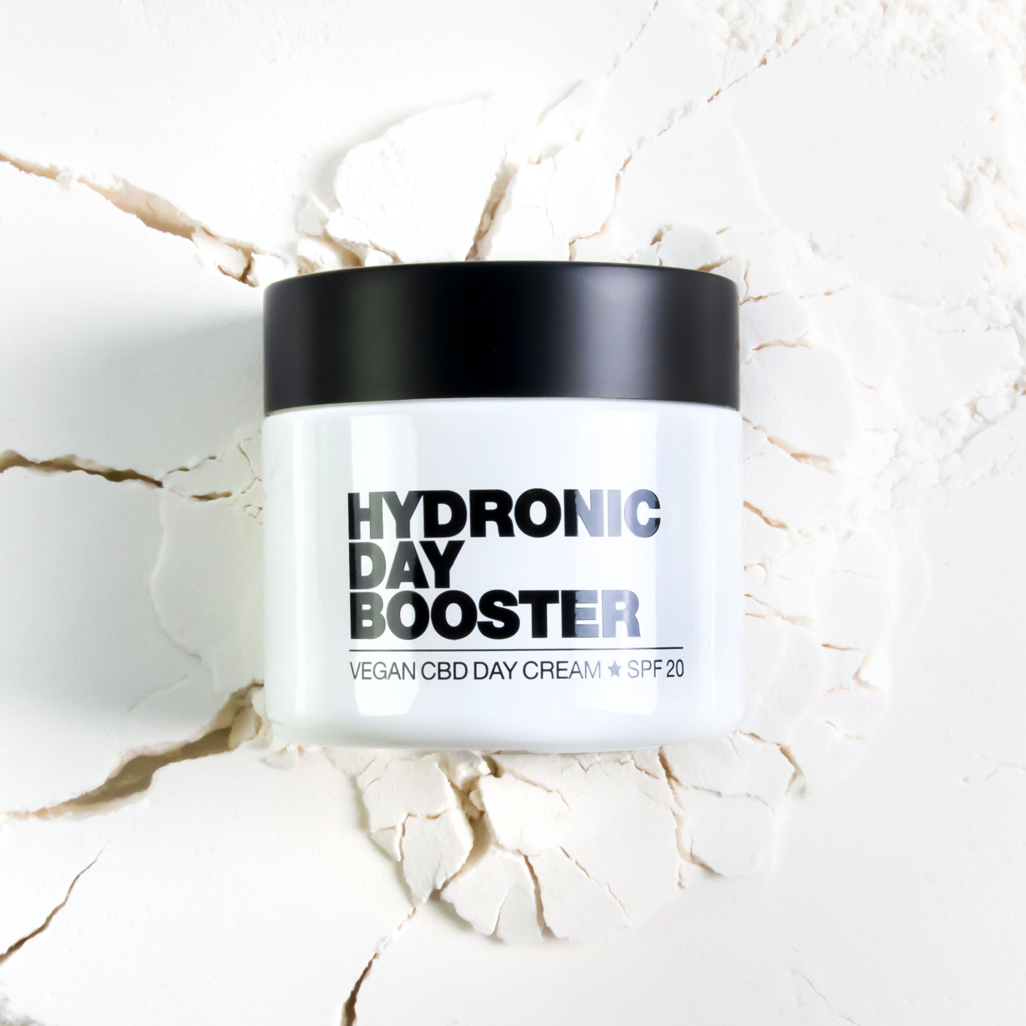 Weißer Tiegel mit schwarzem Deckel. Aufschrift: HYDRONIC DAY BOOSTER, VEGAN CBD DAY CREAM + SPF 20. Liegt auf weißem Pulver.