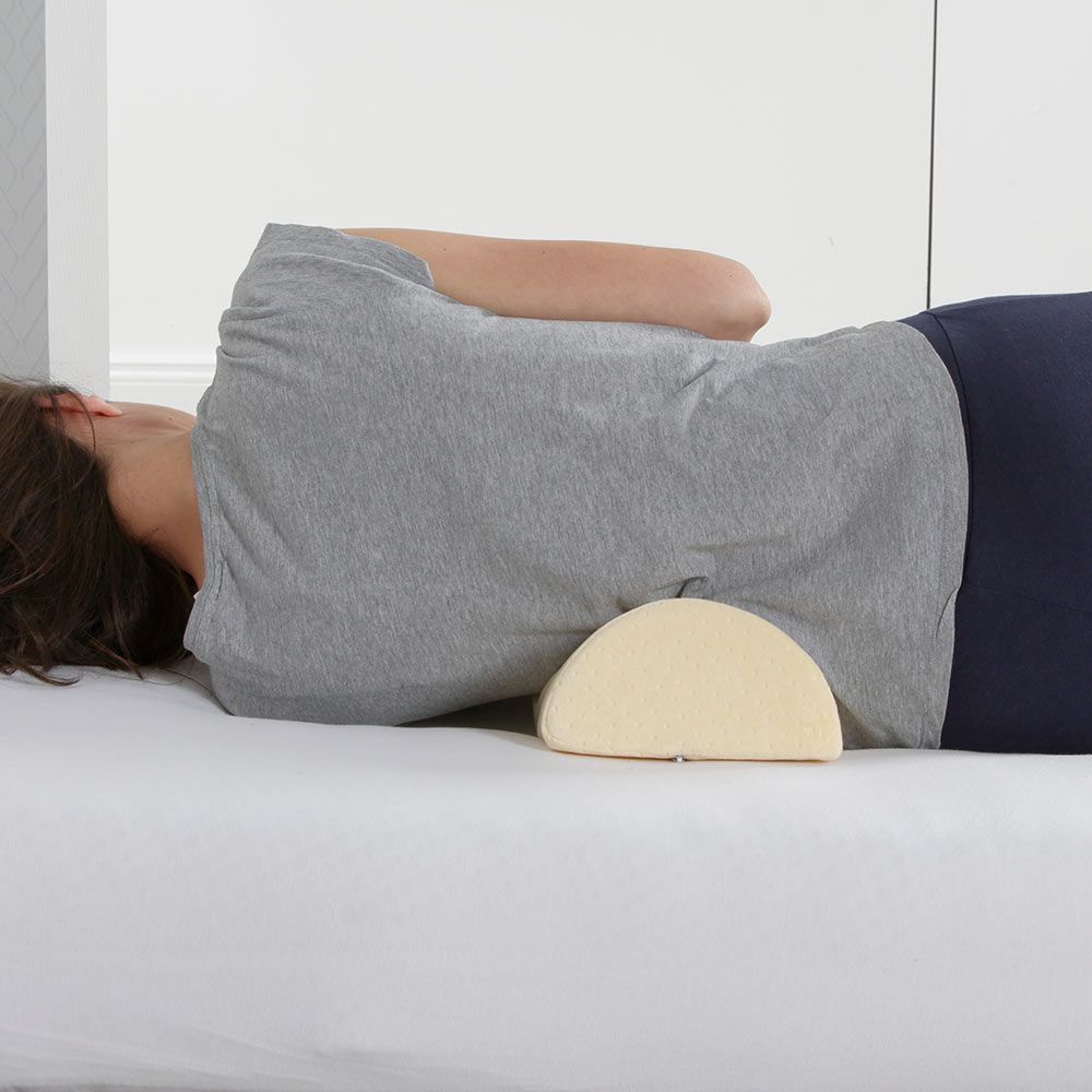 Person liegt auf dem Bauch, Lendenkissen unter dem Rücken. Beige, halbmondförmiges Kissen. Auf weißer Matratze.