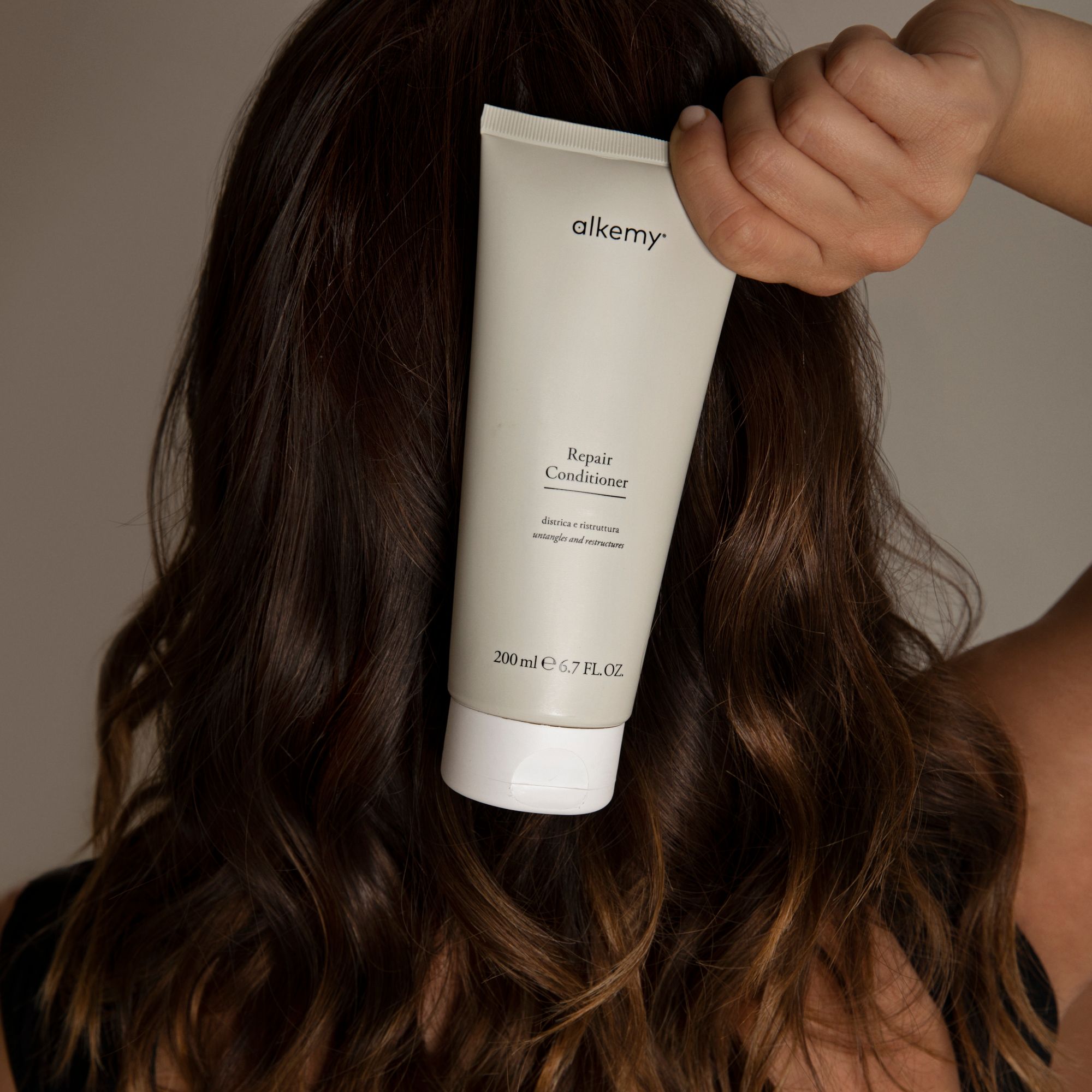 Eine Flasche Alkemy Repair Conditioner, 200 ml, wird hinter dem Haar einer Person gehalten.