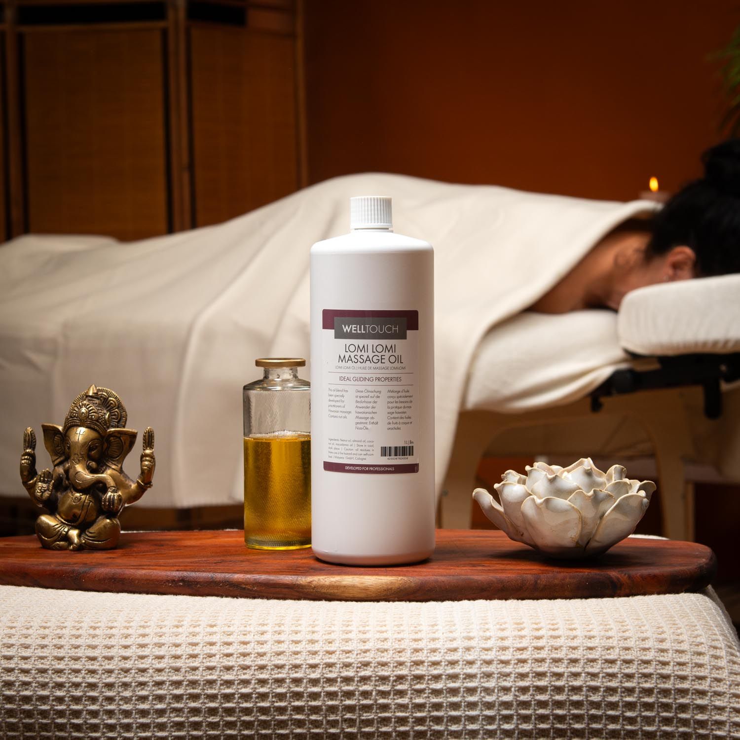 Große weiße Flasche Welltouch Lomi Lomi Massageöl. Daneben eine kleine Glasflasche mit Öl. Im Hintergrund eine Person auf einer Liege.