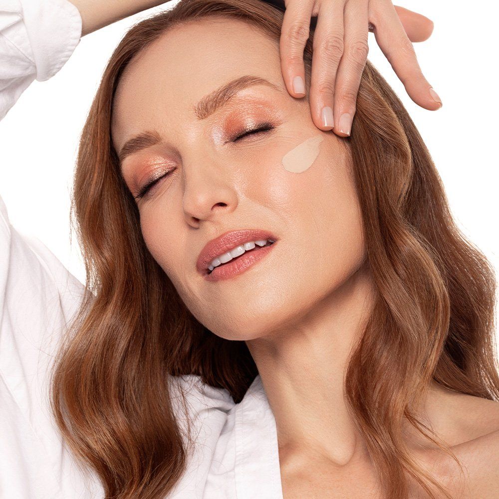 Una donna con capelli rossi ondulati applica il fondotinta sul viso. Una goccia di fondotinta è visibile sulla sua guancia. Ha gli occhi chiusi.