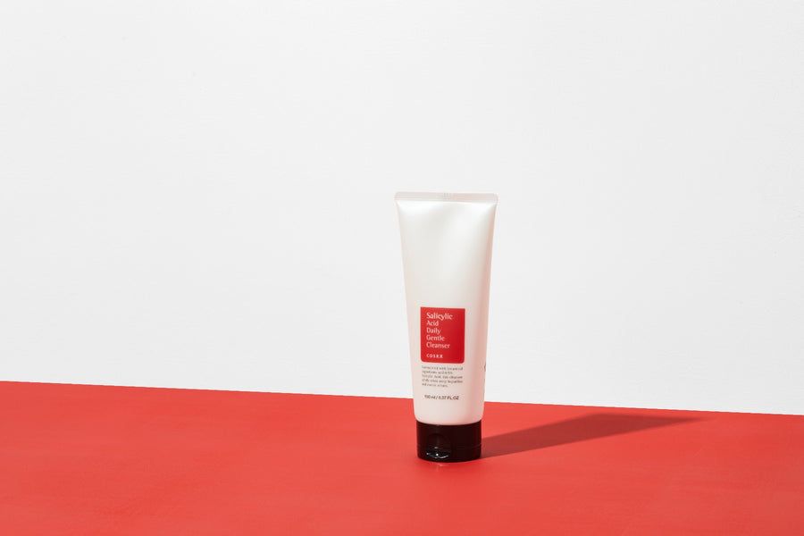 Tubo bianco con etichetta rossa su superficie rossa. Testo: Salicylsäure Daily Gentle Cleanser, COSRX. Tappo nero.