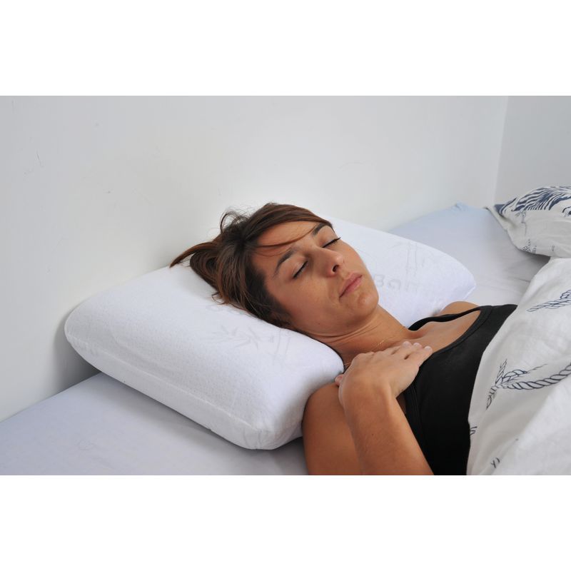 Una donna è sdraiata su un letto, che utilizza un cuscino cervicale ergonomico bianco. Il cuscino ha una forma ondulata e sostiene il collo. È sotto una coperta.