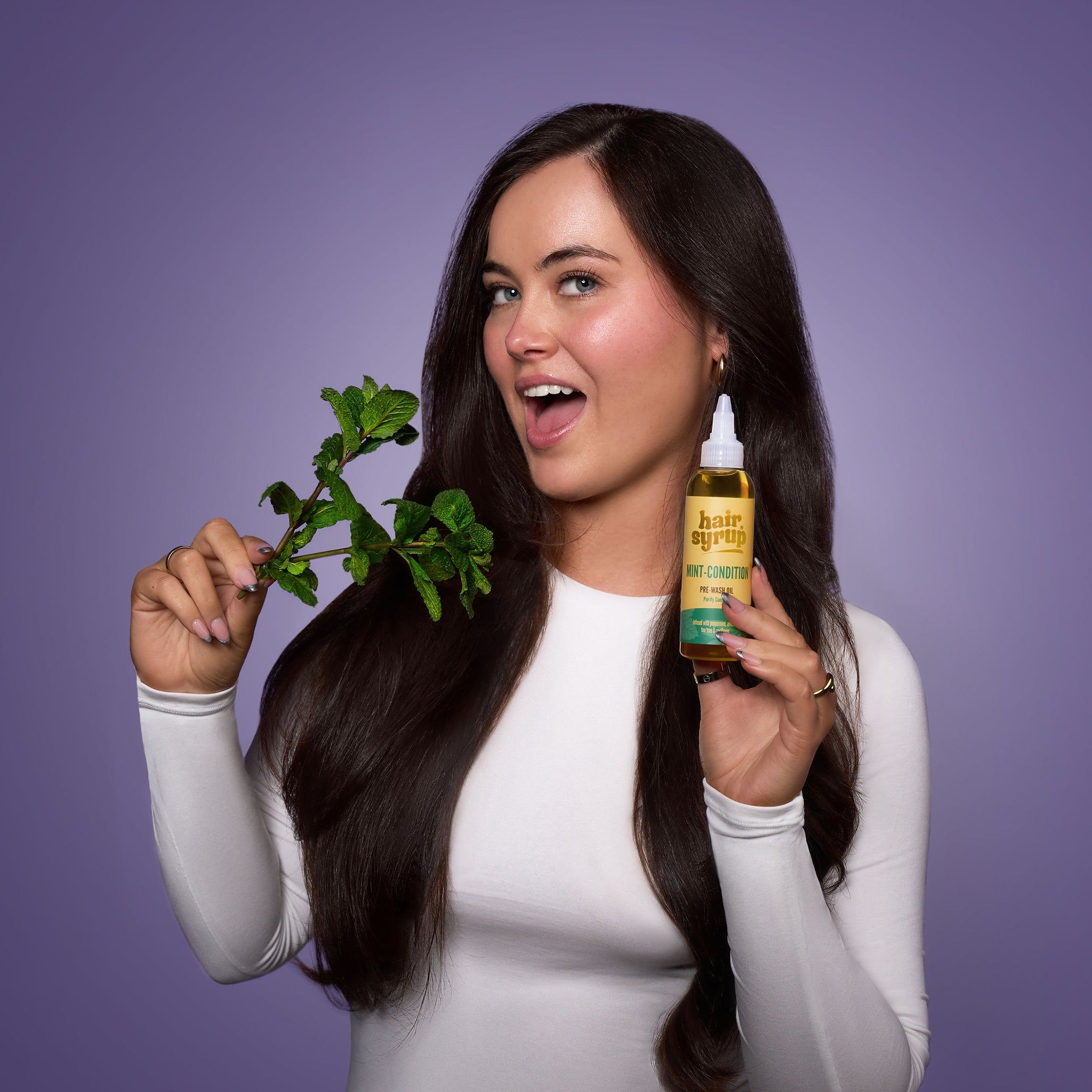 Frau mit langen Haaren hält Flasche "Hair Syrup Mint Condition" und Minzzweig. Lila Hintergrund.