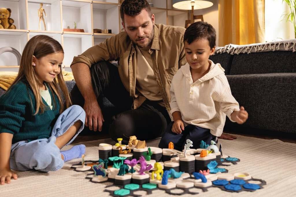 Tre bambini giocano con il Ravensburger GraviTrax Junior Starter-Set XXL Planet. Il gioco è allestito sul pavimento, con vari elementi e personaggi. Un adulto osserva il gioco.