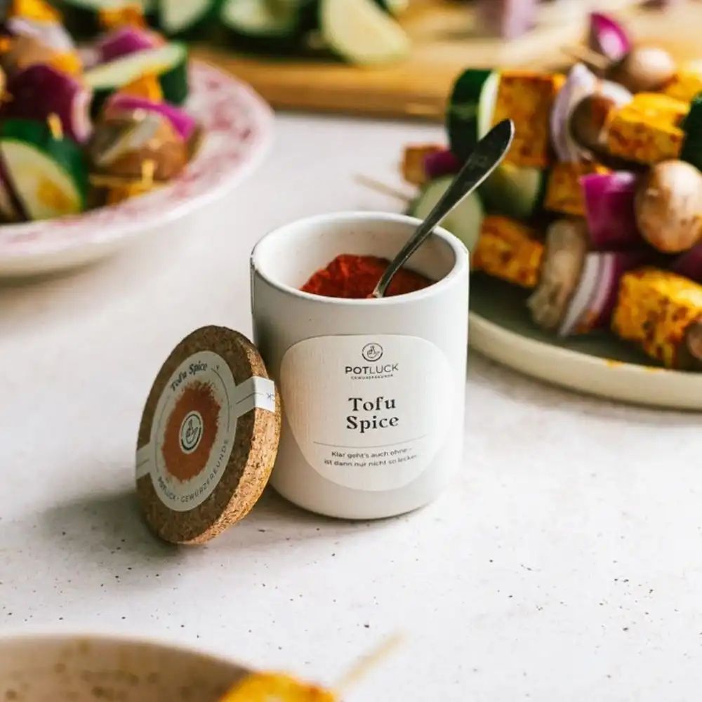 Kleiner weißer Behälter mit Korkdeckel und "POTLUCK Tofu Spice" Etikett. Löffel in Gewürz. Spieße im Hintergrund.