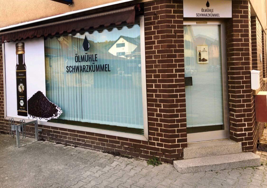 Geschäft mit Fensterwerbung für Schwarzkümmel. Logo: Ölmühle Schwarzkümmel. Außenansicht mit Tür und Werbetafel.