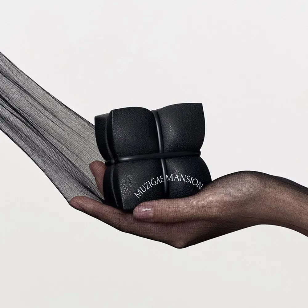 Hand hält schwarzes, quadratisches MUZIGAE MANSION Cushion-Make-up. Schwarzer Stoff im Hintergrund.