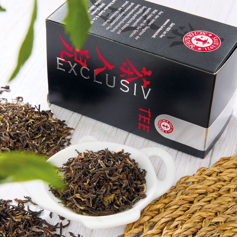 Produktpräsentation mit Tee, Verpackung und Schale. Die Verpackung zeigt "EXCLUSIV" und "TEE". Lose Teeblätter und eine Schale sind ebenfalls zu sehen.