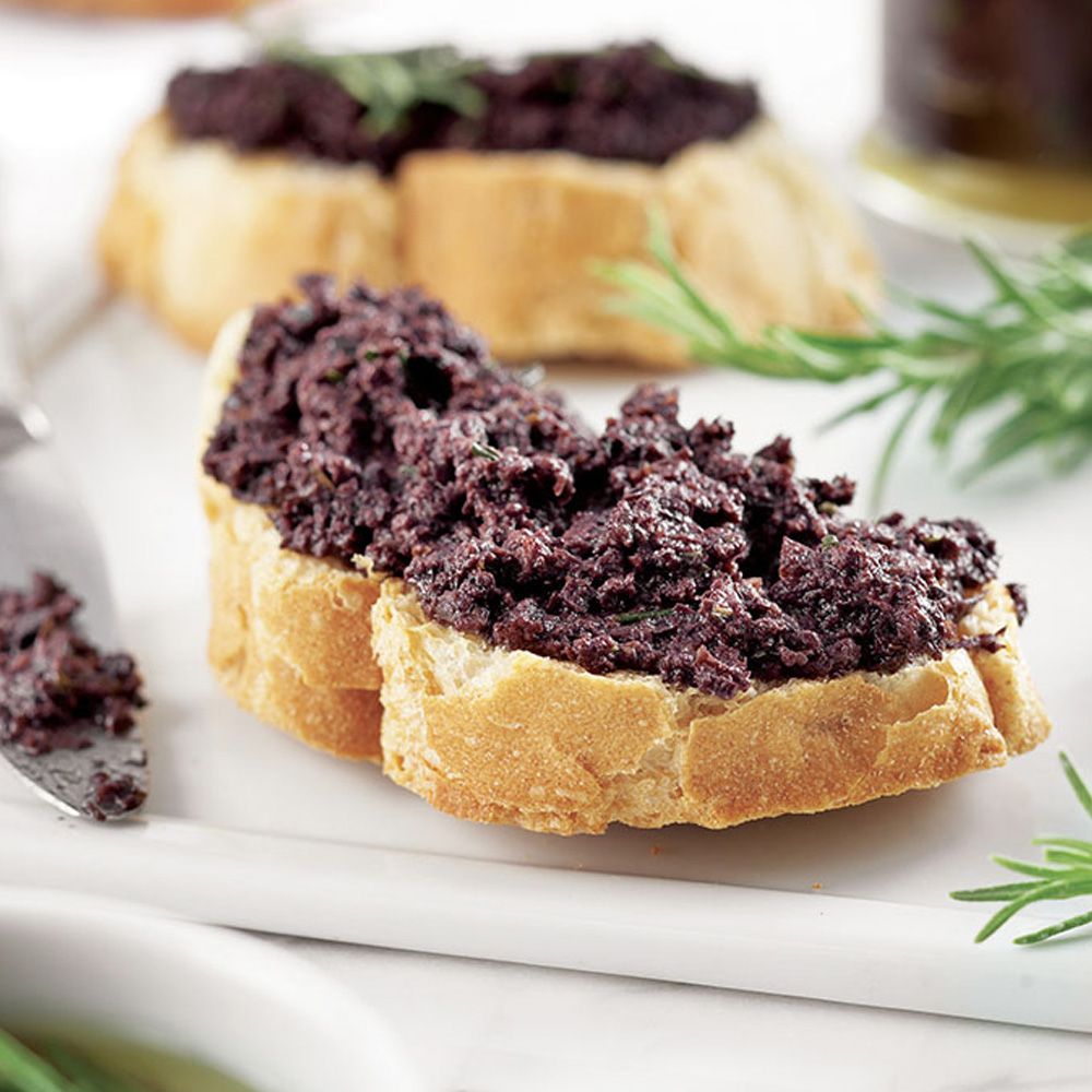 Brotscheiben mit Olivenpaste bestrichen. Rosmarinzweig als Garnitur. Auf weißem Teller.