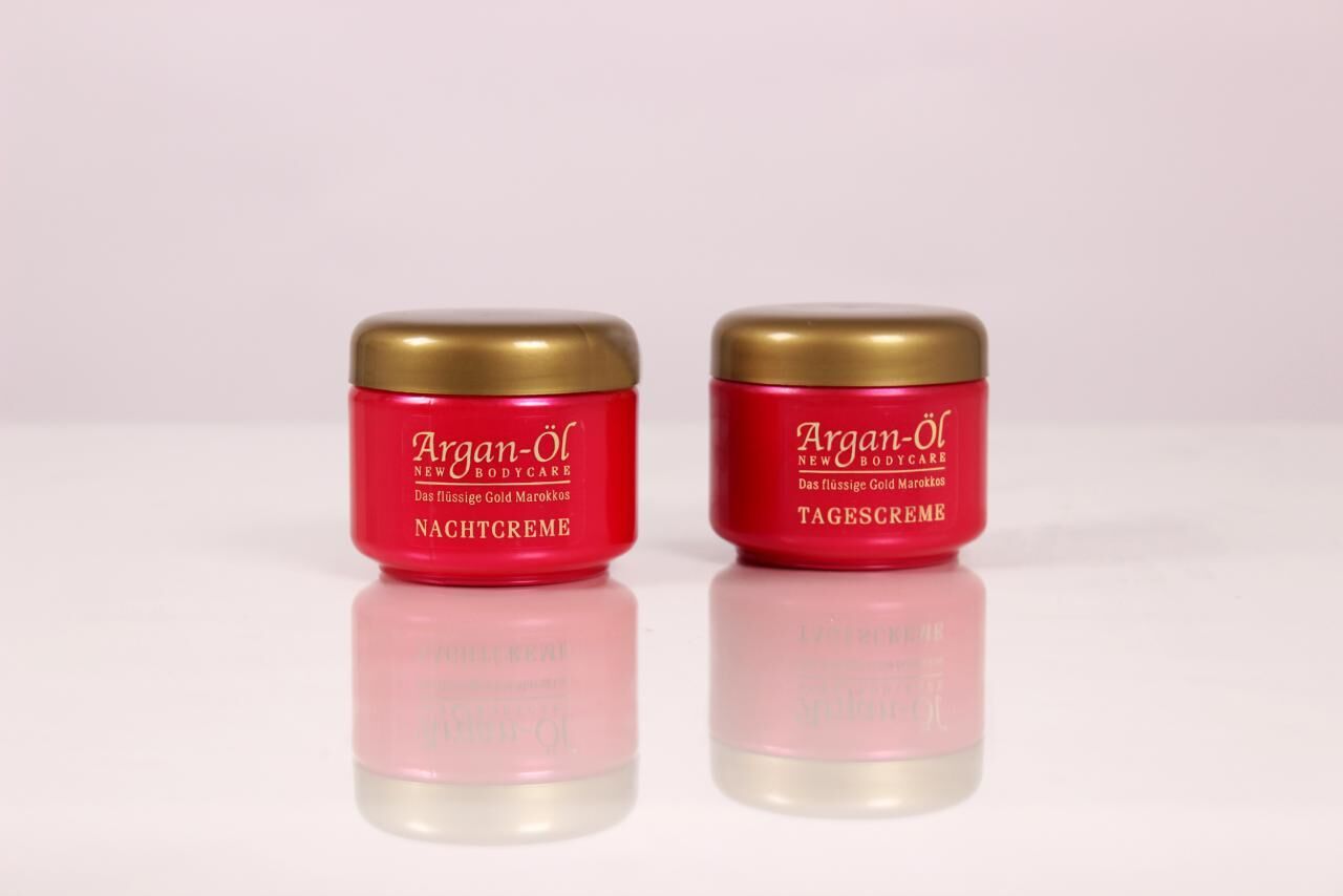 Zwei rote Tiegel mit goldenen Deckeln. Aufschrift: Argan-Öl, Nachtcreme und Tagescreme. Reflektion auf weißem Untergrund.
