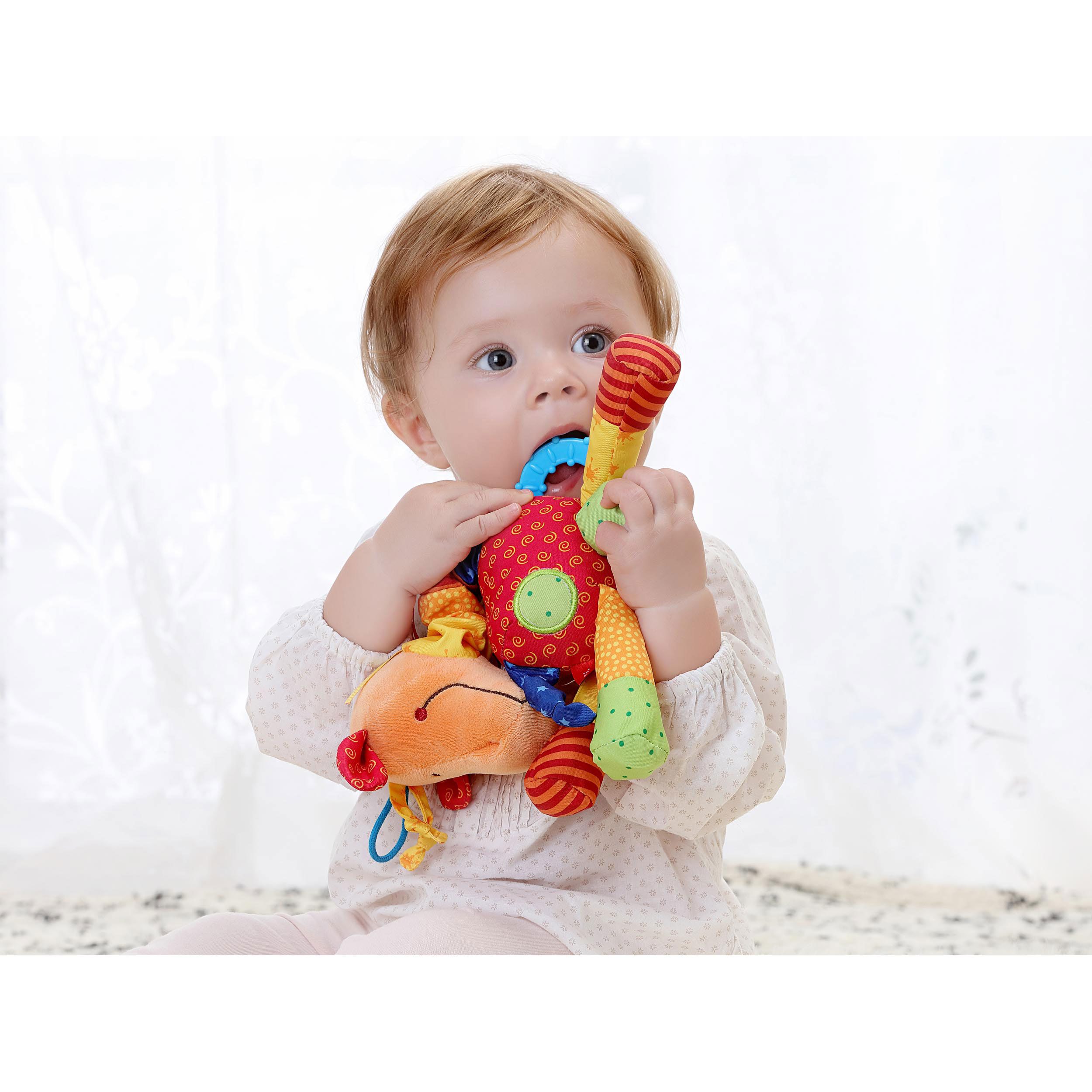 Baby mit Spielfigur im Mund. Orangefarbenes Tier mit bunten Stoffteilen. Blauer Ring.
