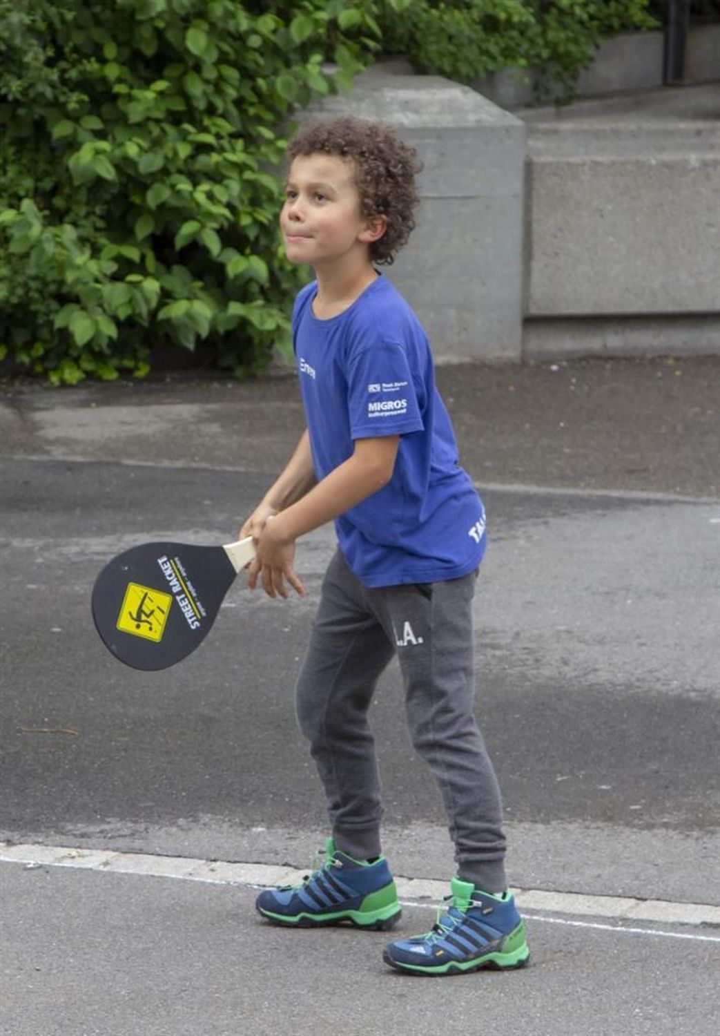 Kind spielt Street Racket. Schwarzer Schläger mit gelbem Logo. Kind trägt blaues T-Shirt und graue Hose. Spielt auf Asphalt.