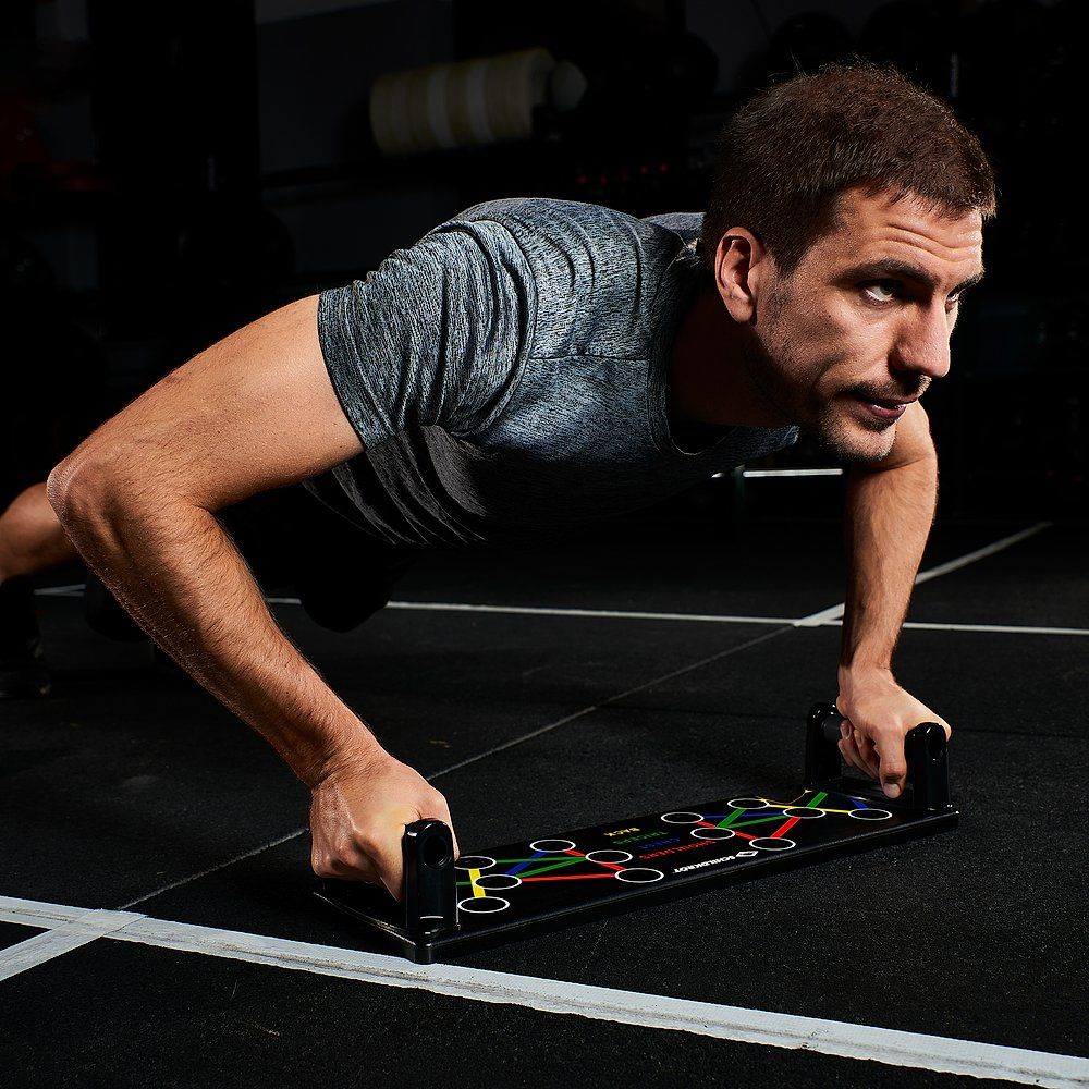 Mann macht Liegestütze auf einem schwarzen Push-Up-Board mit Griffen und farbigen Linien für Muskelgruppen.