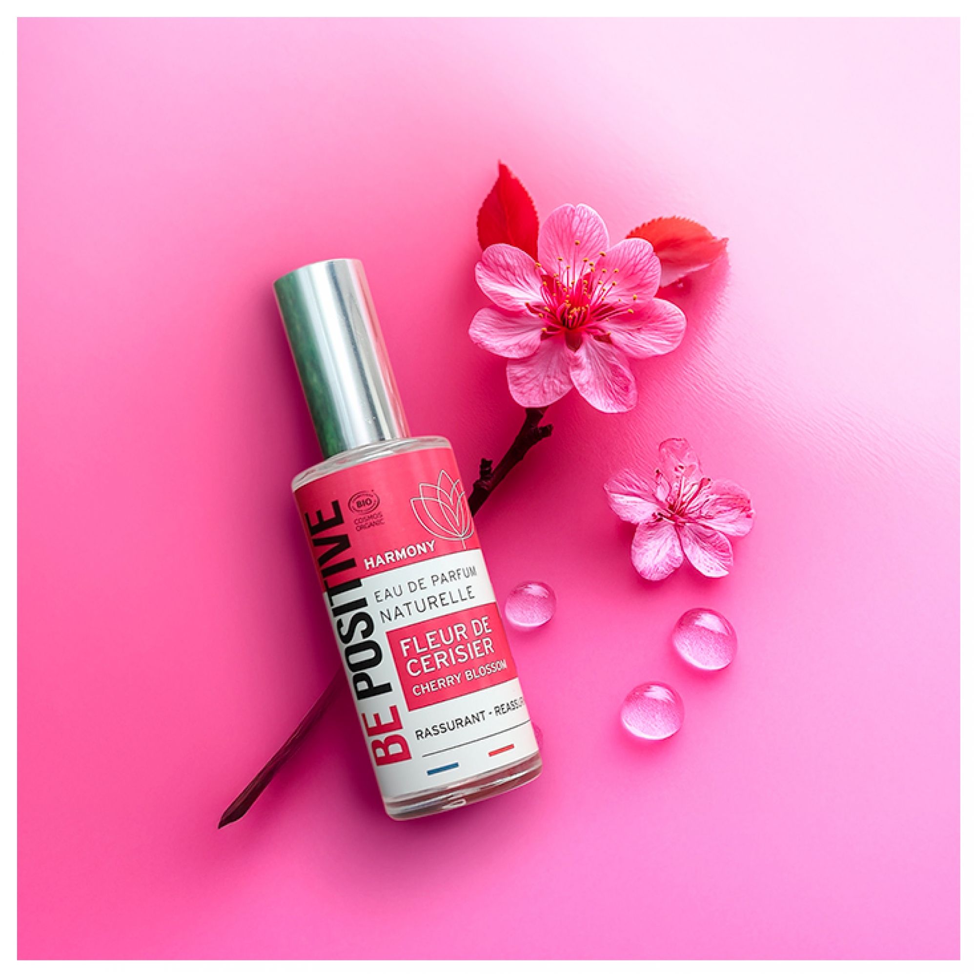 Acorelle Be Positive Eau de Parfum Fleur de Cerisier neben Kirschblüten und Wassertropfen auf pinkem Hintergrund.