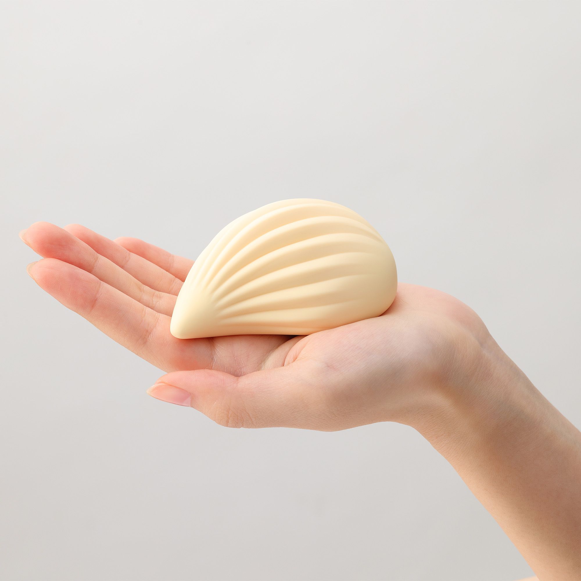 Beigefarbener Vibrator in Tropfenform, gehalten in einer Hand. Geriffelte Oberfläche. Helle Umgebung.