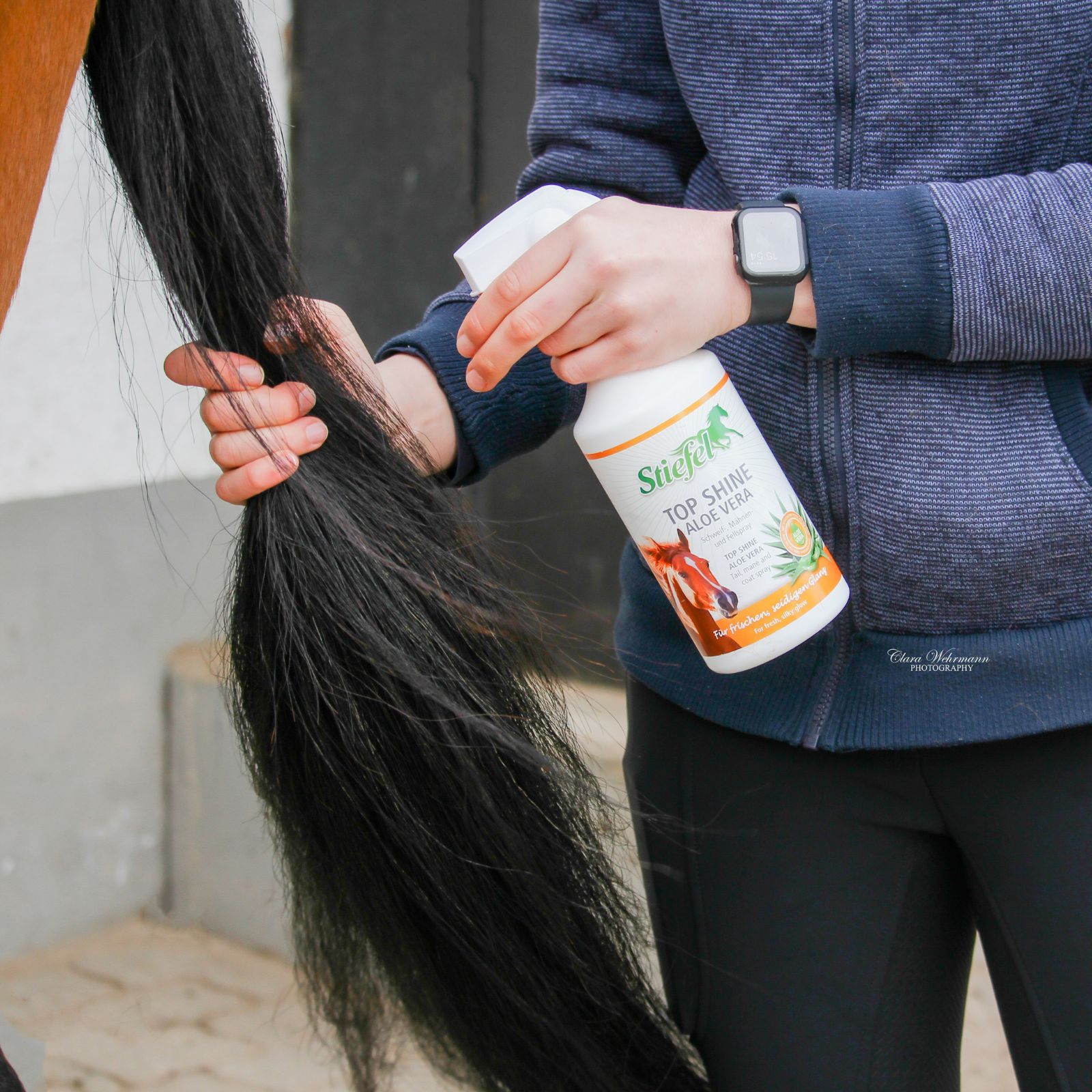 Person sprüht Stiefel Top Shine Aloe Vera auf einen Pferdeschwanz. Weiße Flasche mit Sprühkopf.