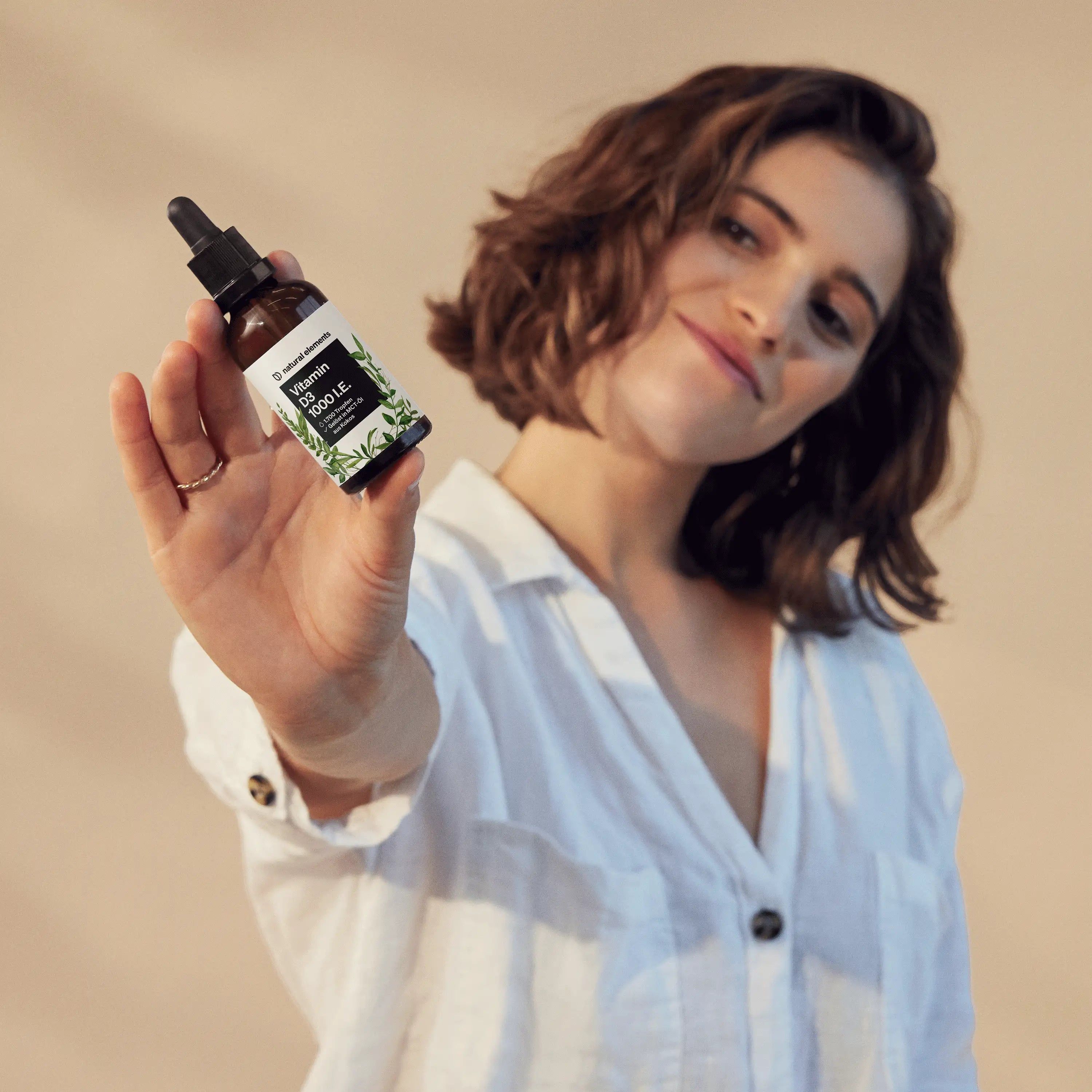 Frau hält eine braune Flasche mit "Vitamin D3 1000 I.E." in die Kamera. Sie trägt ein weißes Hemd.