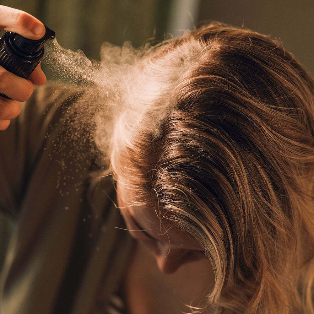 Nahaufnahme einer Person, die Trockenshampoo auf ihr Haar sprüht. Das Produkt ist nicht sichtbar.