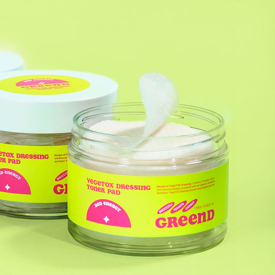 Offener Glasbehälter mit gelbem Etikett. Aufschrift: Vegitox Dressing Toner Pad, GreenD. Rosa Logo. Pad wird entnommen.