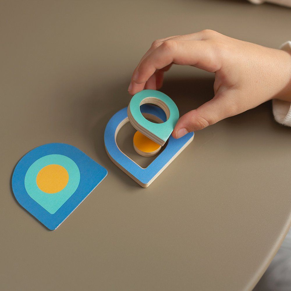 Kind greift nach einem blauen Tropfen-Form-Puzzleteil mit einem grünen Kreis. Ein weiteres Puzzleteil liegt daneben.