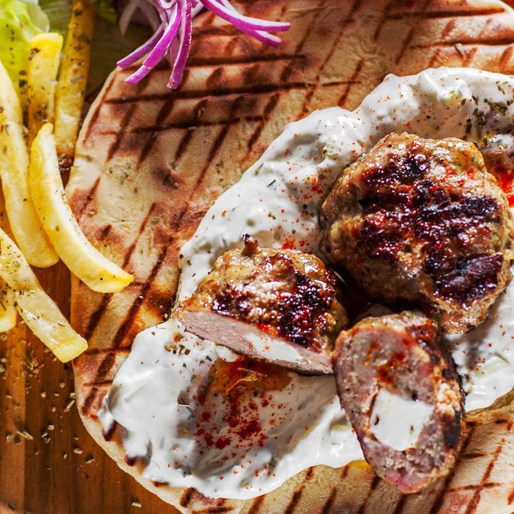 Gegrillte Frikadellen auf Fladenbrot mit Sauce und Pommes. Einige Frikadellen sind aufgeschnitten.