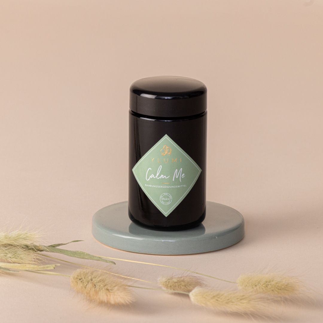 Schwarze Dose mit grünem Etikett. Aufschrift "Calm Me". Steht auf einem grauen Podest, umgeben von dekorativen Gräsern.