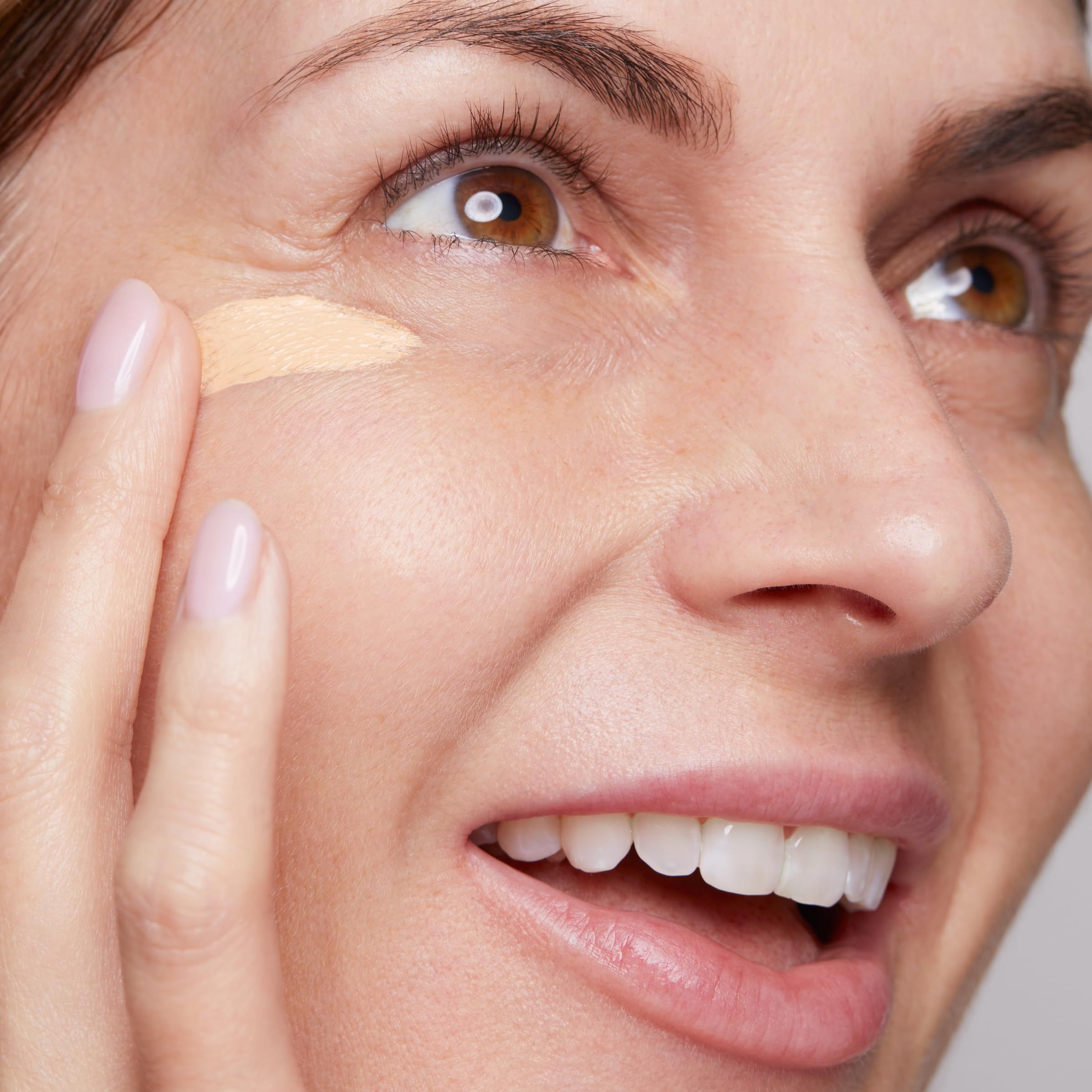 Frau trägt Primer auf. Beige Flüssigkeit auf der Haut. Sie lächelt.