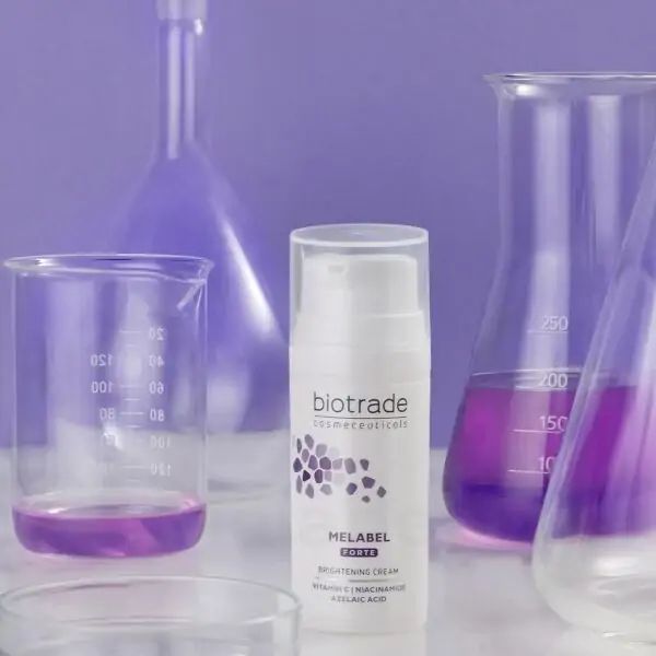 Weiße Flasche mit Sprühkopf, umgeben von Laborgeräten. Aufschrift: Biotrade, Melabel Forte, Brightening Cream. Enthält Vitamin C, Niacinamid, Azelainsäure.