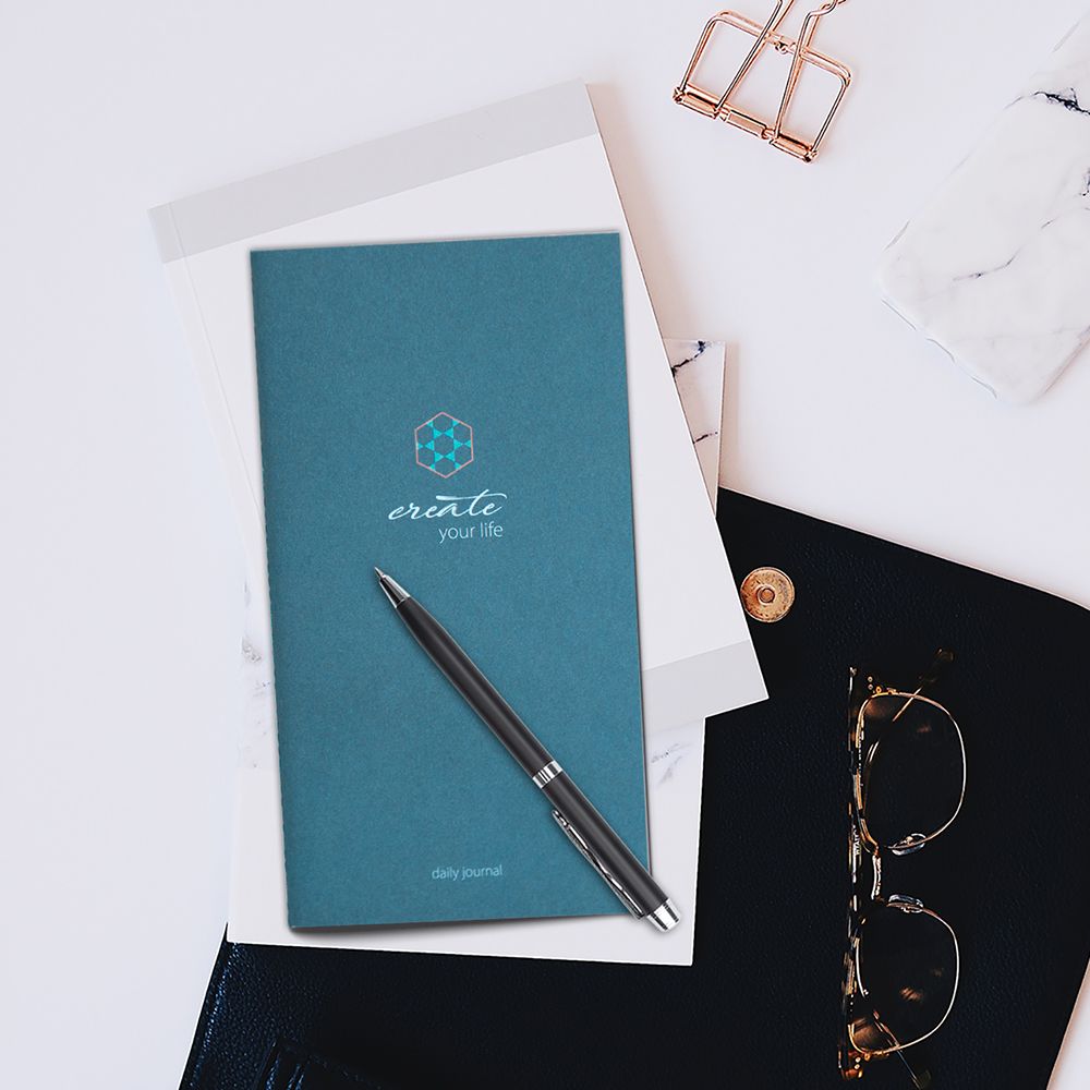 Blaues Tagebuch mit Stift auf weißem Untergrund. Aufschrift: create your life, daily journal. Goldene Büroklammer.