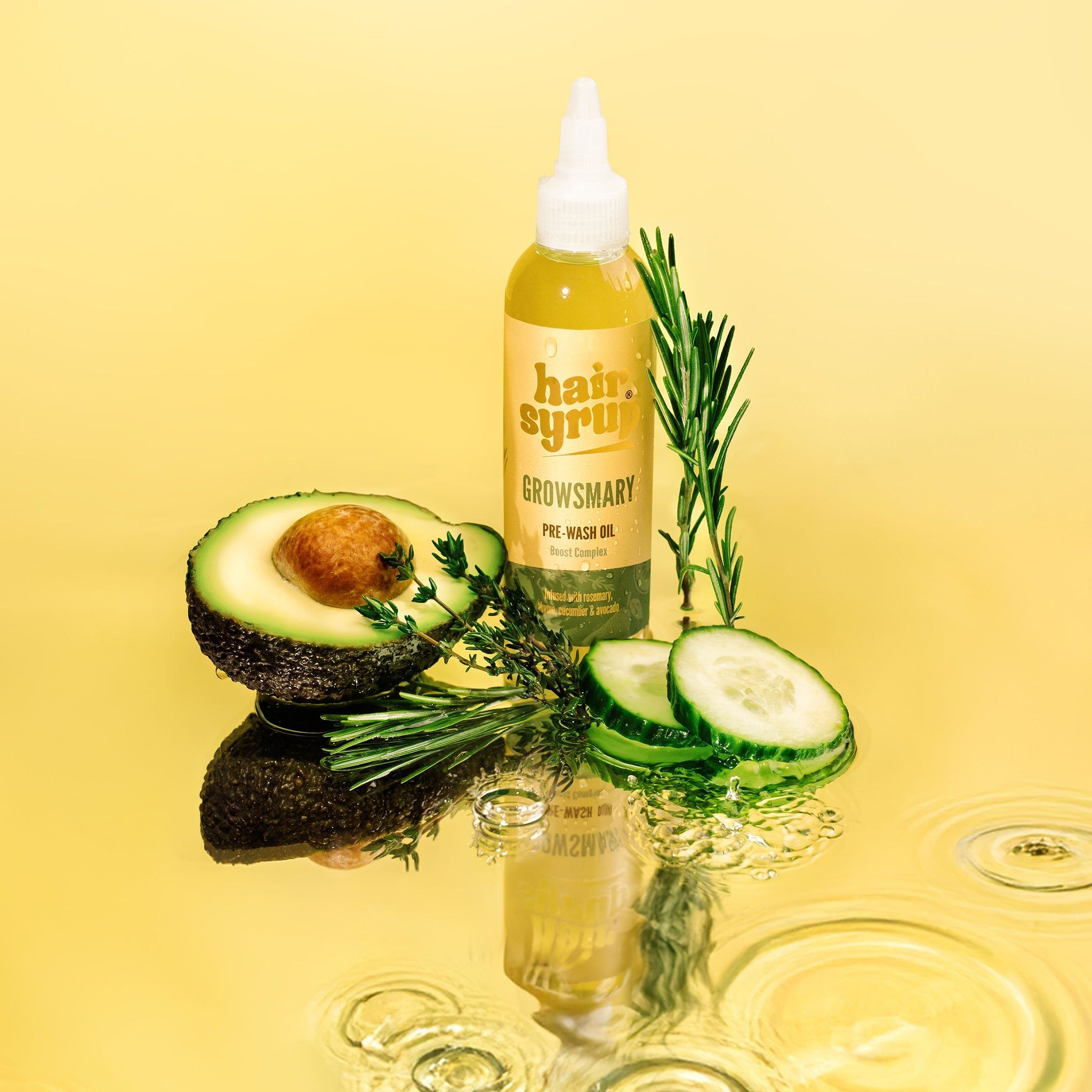 Gelbe Flasche mit Produktetikett, daneben Avocado, Gurke, Rosmarin. Reflektierende Oberfläche.