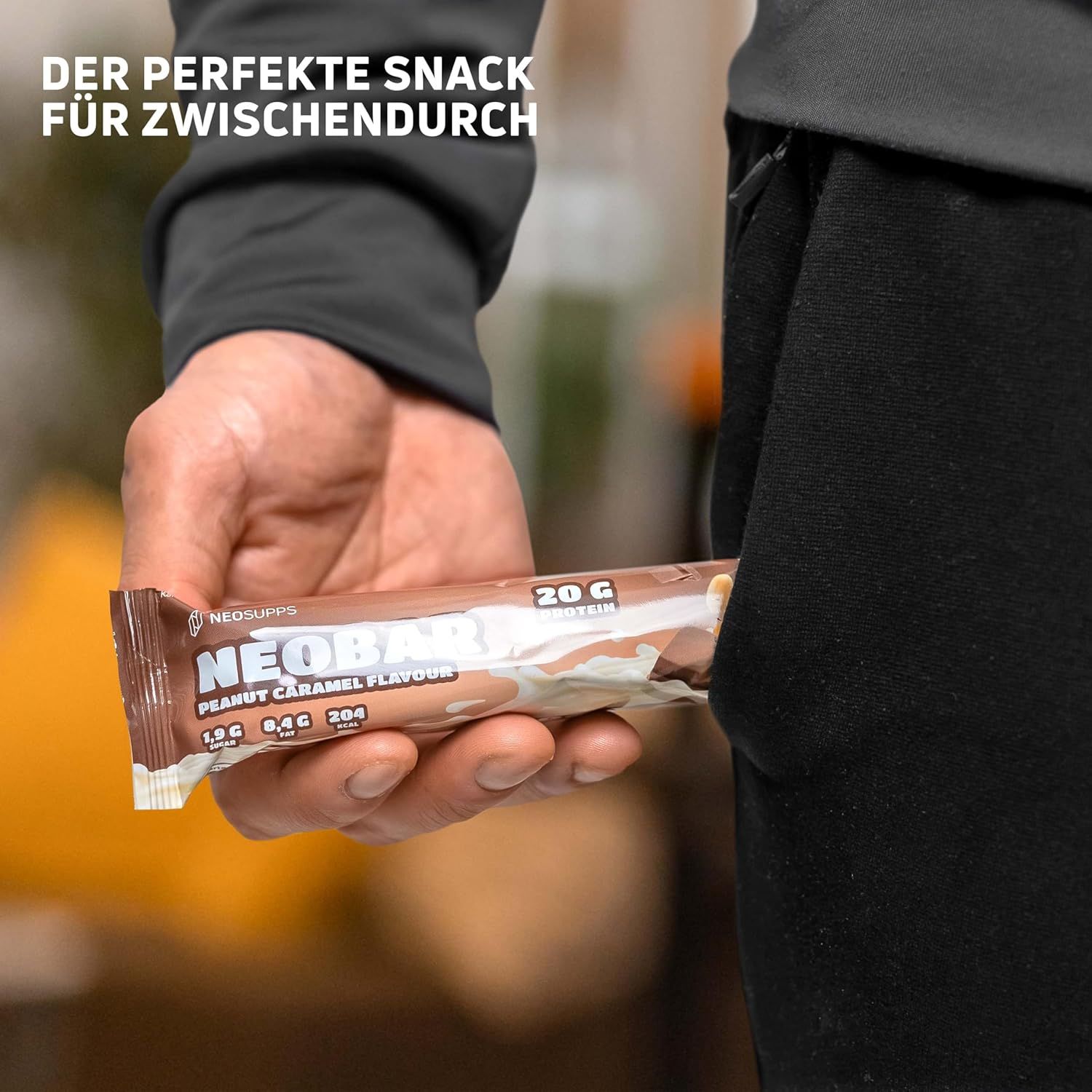 Person hält NeoBar-Riegel in der Hand. Aufschrift: NeoBar Peanut Caramel Flavour, 20g Protein. Riegel in der Hosentasche.