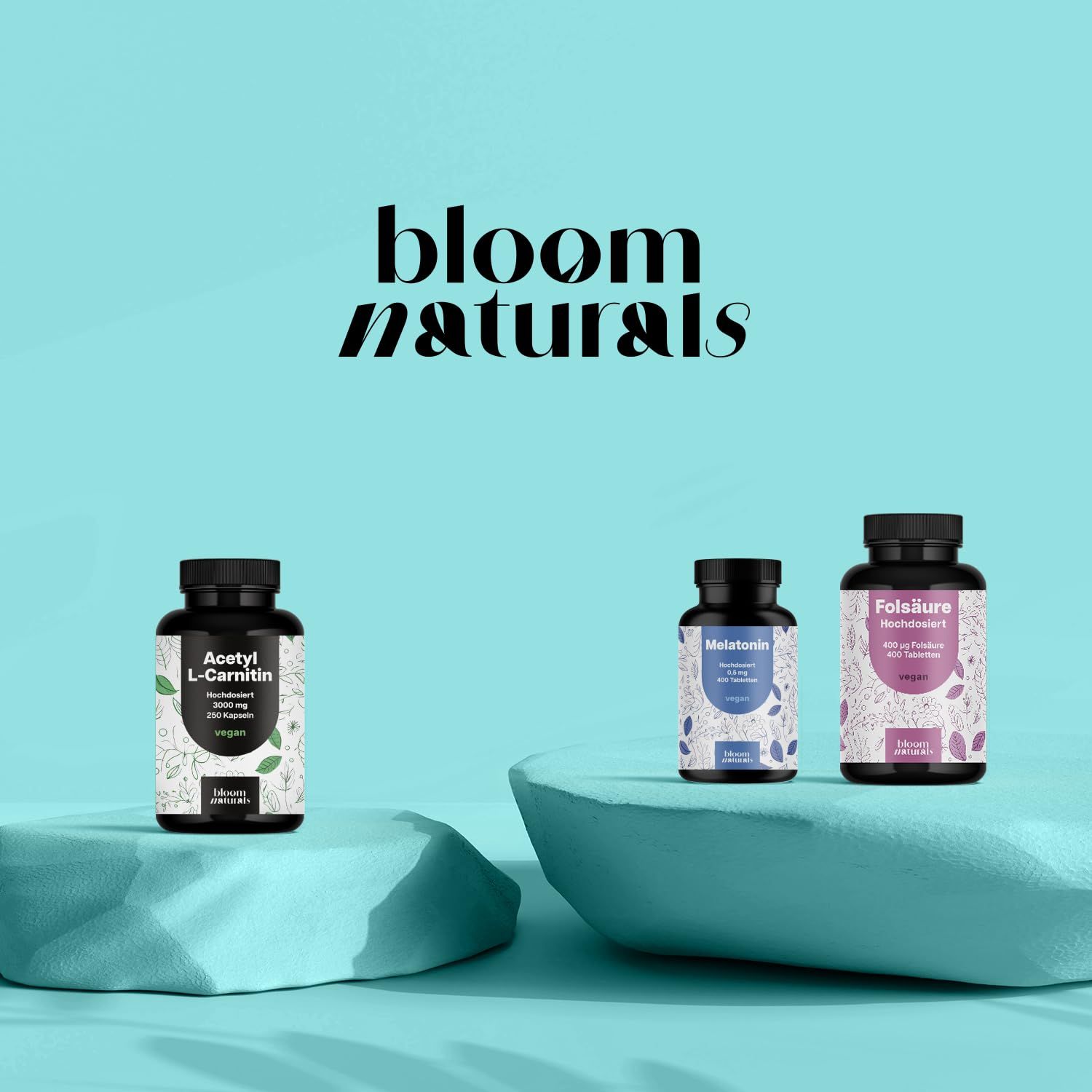 Drei Bloom Naturals Flaschen: Acetyl-L-Carnitin, Melatonin, Folsäure. Auf Podesten.