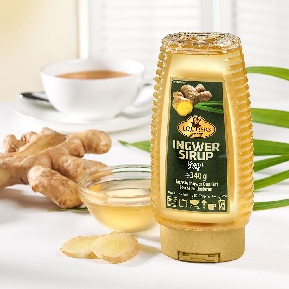 Ingwersirup-Flasche neben Ingwerstücken, Schale mit Sirup und Tasse Tee. Etikett: Ingwer Sirup Vegan.