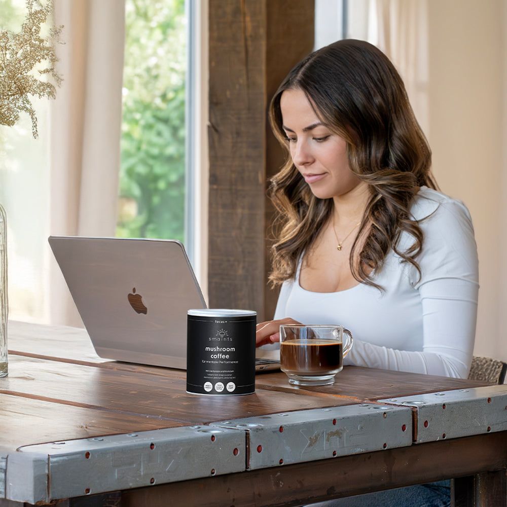 Frau am Laptop mit Dose und Tasse Kaffee auf Holztisch. Dose mit Text: Mushroom Coffee, für mentale Performance.