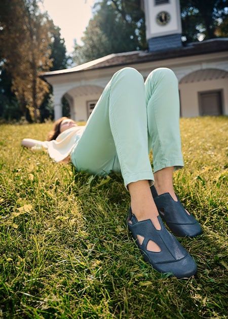 Frau liegt auf Rasen, trägt blaue Sandalen mit breiter Passform. Offene Zehenpartie, Fersenriemen. Hintergrund: Gebäude.