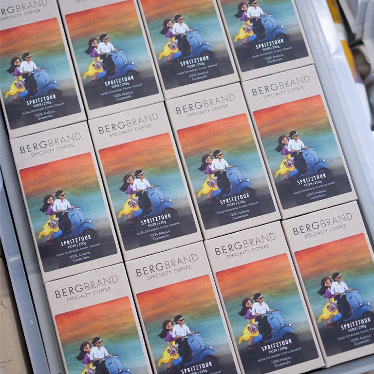 Mehrere Packungen BERGBRAND Spritztour Kaffee. Kartons mit Illustration von Personen auf einem Roller.