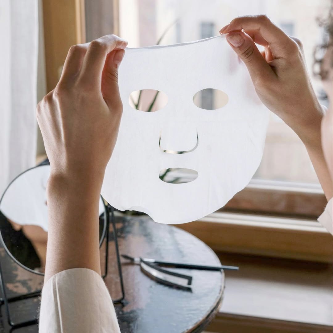 Hände halten eine weiße, entfaltete Alkemy Gesichtsmaske mit Augen- und Mundöffnungen.