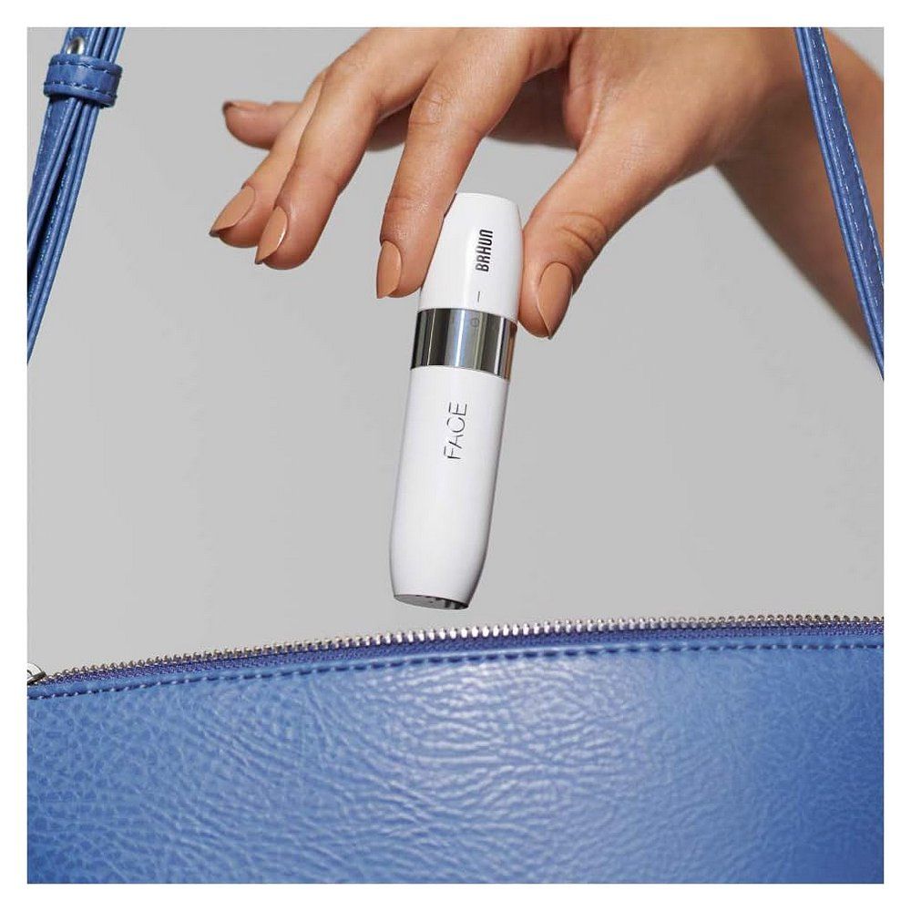 Hand hält einen weißen elektrischen Gesichtsrasierer über einer blauen Handtasche.